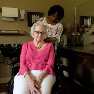 An elderly woman in a wheelchair smiles as a caregiver styles her hair in a cozy indoor setting.
