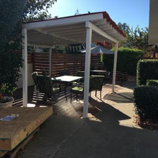 A cozy outdoor patio area featuring a shaded pergola with a table and chairs, surrounded by greenery.