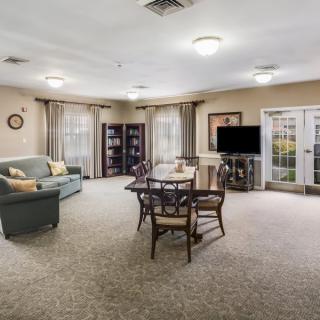 A spacious living room with a cozy sofa, dining table, bookshelf, and television, featuring carpeted flooring and ample natural light.