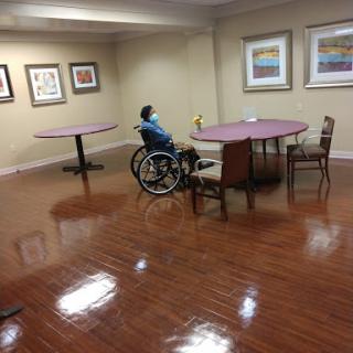 A spacious indoor area featuring a person in a wheelchair, surrounded by tables and chairs, with art on the walls.