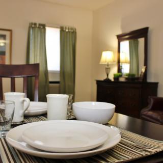 A warmly lit dining room with neatly arranged tableware on a table, featuring plates, bowls, and cups. The room includes wooden furniture and green curtains.