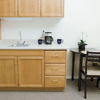 A small kitchen area with wooden cabinets, a sink, a coffee maker, and mugs on the countertop, accompanied by a table with chairs and a potted plant.
