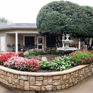 A beautifully landscaped garden with vibrant flowers, a stone wall, and a fountain in front of a residential home.