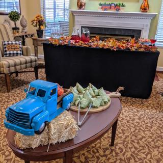 An indoor autumn-themed display featuring a blue miniature truck with pumpkins on a hay bale and a fireplace adorned with fall decorations.