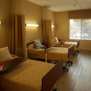 A shared healthcare room with two beds, separated by curtains and lit by wall-mounted lights.