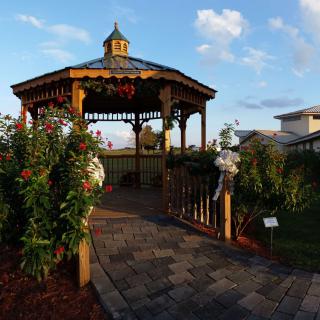 A charming wooden gazebo in a garden setting with blooming flowers and a small bridge.