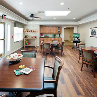 A well-lit community room with wooden flooring, equipped with tables and chairs, a ceiling fan, and ample natural light pouring in from large windows.