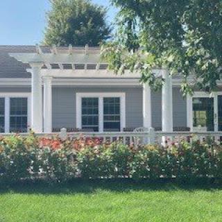 A cozy house with a white pergola over the porch, surrounded by green hedges and a neatly maintained lawn.
