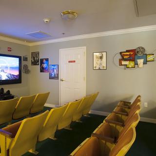 A small indoor theater room with rows of yellow chairs facing a large screen, adorned with movie posters on the walls.
