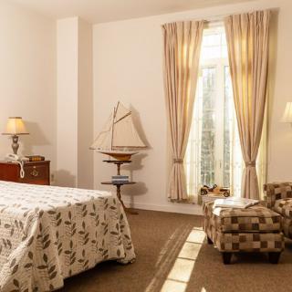 A cozy bedroom interior featuring a floral bedspread, patterned armchair, and elegant curtains with light streaming through the window.