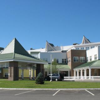 A modern building with a green roof and a large entrance, possibly a hospital, under a clear blue sky.
