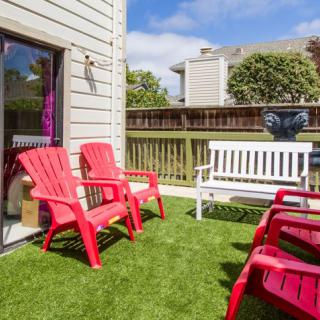 A cozy outdoor patio featuring red chairs and a white bench on an artificial grass surface, surrounded by a wooden fence under a clear blue sky.