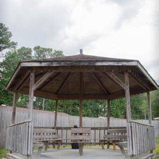 A wooden gazebo with benches surrounded by a fence and trees under a cloudy sky.