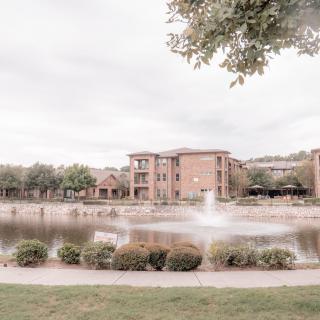 A serene view of a lake with a fountain, surrounded by buildings and trees under an overcast sky.