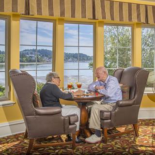 Two seniors enjoying a meal with a scenic lake view through large windows.