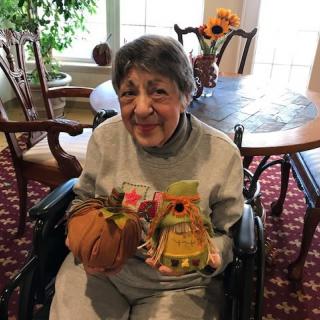 An elderly woman in a wheelchair smiles while holding crafts and a small pumpkin indoors.
