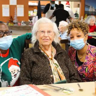 Three women, two wearing masks, gather together at a community event.