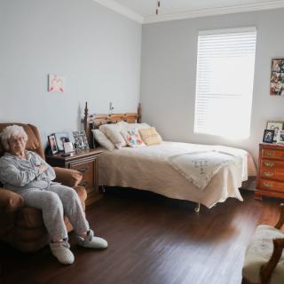 An elderly person sitting in a comfortable chair in a cozy, well-lit bedroom.
