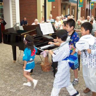 Children in kimonos playing near a piano at an outdoor festival.
