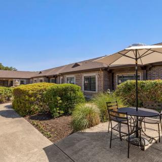 A patio area with chairs and an umbrella, surrounded by greenery and a building in the background.