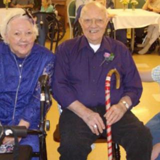 An elderly couple seated indoors; the woman is in a wheelchair, and the man is holding a cane.