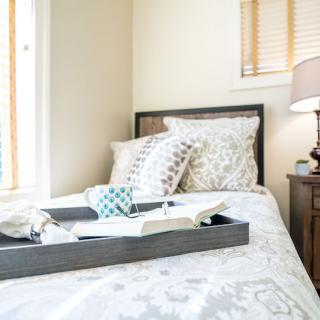 A cozy bedroom with a neatly arranged bed, featuring patterned pillows and a decorative tray alongside a bedside lamp.