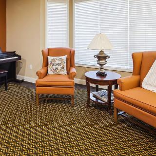 A cozy living room with orange armchairs, a matching sofa, a piano in the corner, patterned carpet flooring, and window blinds.