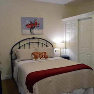 A cozy bedroom with a bed adorned with pillows and a red throw blanket, a lamp on the nightstand, and a floral painting on the wall.