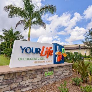 A sign for 'Your Life of Coconut Creek Memory Care' surrounded by palm trees and landscaping.