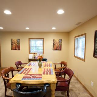 A cozy dining room with a wooden table and chairs, surrounded by artwork and windows providing natural light.