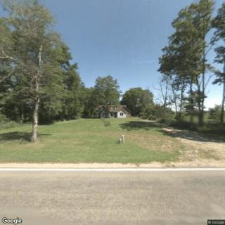 A small house surrounded by trees, situated on a grassy lot with a road in the foreground.
