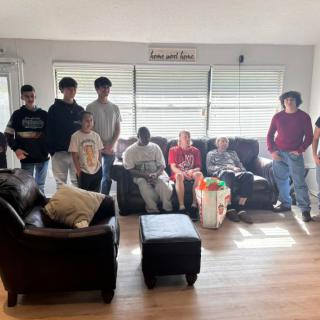 A group of people gathered in a living room, sitting and standing around sofas in a casual setting.