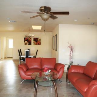 A modern living room with red sofas, a glass coffee table, and a ceiling fan, leading into a dining area.