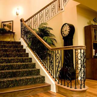 A warmly lit interior featuring a carpeted staircase with ornate railings, a grandfather clock, and decorative elements such as a plant and bookshelf.