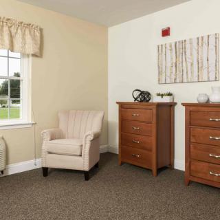 A cozy living room corner featuring an armchair, two wooden dressers, and a window with curtains.