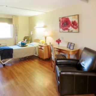 A cozy hospital room with a bed, chair, desk, and bright window.