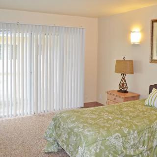 A cozy bedroom featuring a bed with a green patterned cover, a nightstand with a lamp, and vertical blinds covering the window.