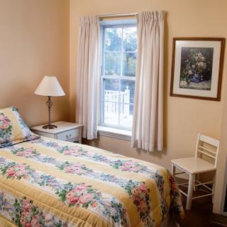 A cozy bedroom with a single bed featuring floral bedding, a lamp on the bedside table, and a window with light curtains.