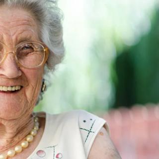 An elderly woman smiling warmly outdoors, wearing glasses and a pearl necklace.
