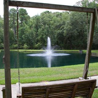 A peaceful scene with a wooden swing overlooking a pond and water fountain surrounded by trees.