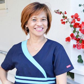 A woman in nursing scrubs smiling outdoors near vibrant flowers.