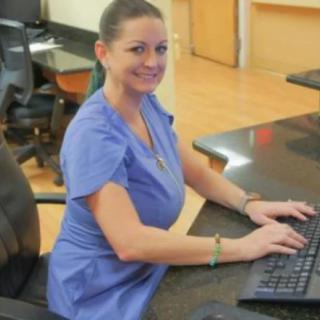 A nurse in uniform working at a desk with a computer, smiling at the camera.