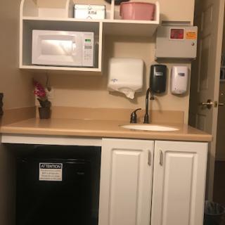 A small kitchenette area featuring a microwave, sink, and refrigerator with shelving for storage.