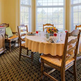 A cozy dining room with a round table set for a meal, surrounded by wooden chairs and large windows letting in natural light.