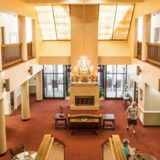 A spacious lobby area with a chandelier, carpeted flooring, and various furniture pieces, including tables and chairs. People are seen walking around the space.