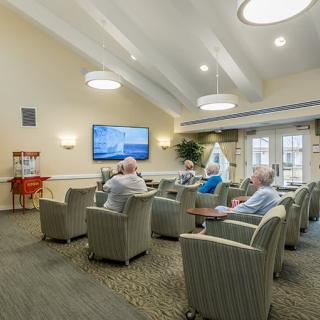 A group of seniors sitting in a community room watching television and socializing.