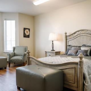 A cozy bedroom featuring a large bed with a decorative headboard, accompanied by two chairs and a lamp, bathed in natural light from a tall window.