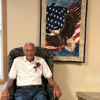 An elderly man sitting in an armchair next to a patriotic quilt featuring an eagle and the American flag.