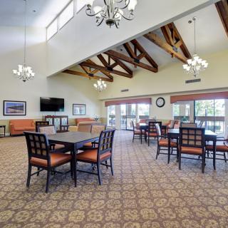 A spacious dining area with multiple tables and chairs, featuring chandeliers and exposed wooden beams under a high ceiling.