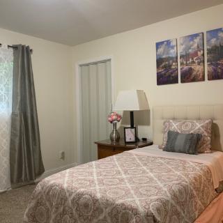 A cozy bedroom with a single bed, adorned with patterned bedding and a decorative throw pillow. The room features curtains, a bedside table with a lamp, floral arrangement, and wall art above the headboard.